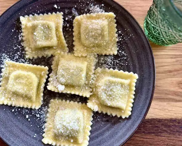 Pięć kwadratowych ravioli posypanych parmezanem na ciemnym talerzu. Idealne do domowego ravioli przepis.