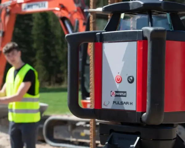 Niwelator laserowy Pulsar H na budowie. W tle pracownik w kamizelce odblaskowej i koparka.