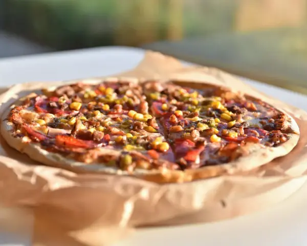 Apetyczna pizza Giuseppe 4 sery, z kukurydzą i mięsem, na papierze do pieczenia, w słonecznym świetle.