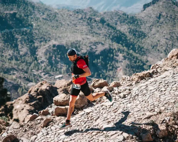Un corredor de trail desciende por un sendero rocoso y empinado, con montañas boscosas al fondo.