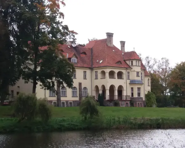 Malowniczy pałac z czerwoną, dachówkową, spadzistą dachem, otoczony zielenią i wodą.