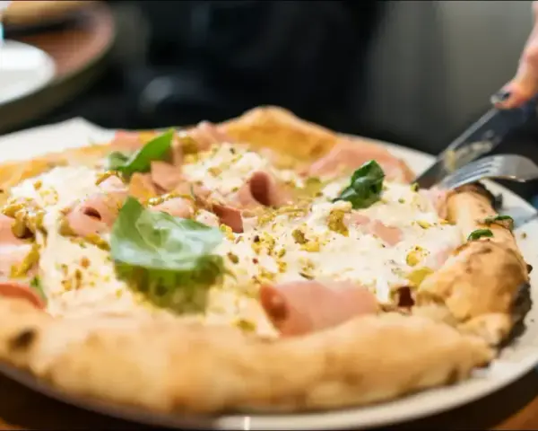 En person skär i en pizza bianco med skinka och basilika. Ett glas vatten står bredvid. Perfekt vin till pizza bianco.