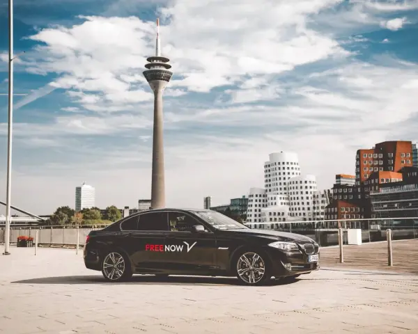 Schwarzer Wagen mit FREE NOW Logo steht vor dem Rheinturm in Düsseldorf. Ein FREE NOW Fahrer wartet auf seine Fahrgäste.
