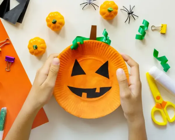 Barn gör halloween pyssel med en pumpa av papper. Små pumpor, spindlar och glitterpennor ligger redo.