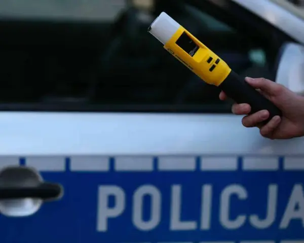 Dłoń trzymająca żółty alkomat na tle radiowozu policyjnego z napisem "POLICJA".