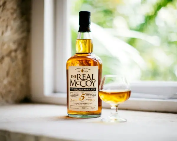 Ein besonderer Rum, "The Real McCoy", steht neben einem Glas mit bernsteinfarbener Flüssigkeit vor einem Fenster mit grünem Bokeh.