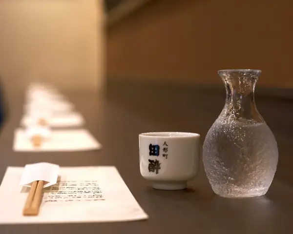 Sake ile procent? Delikatna czarka i karafka z sake czekają na degustację. W tle rząd nakryć.