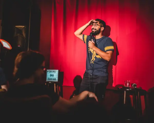 Ein Comedian steht auf einer Bühne in Berlin, hält ein Mikrofon und wischt sich die Stirn. Ein Schild mit "Chips und Kaviar" steht daneben.
