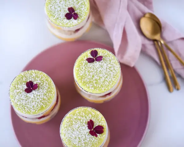 Fyra små prinsesstårtor i glas, dekorerade med florsocker och en lila blomma. En rosa duk och guldfärgade skedar ligger bredvid.