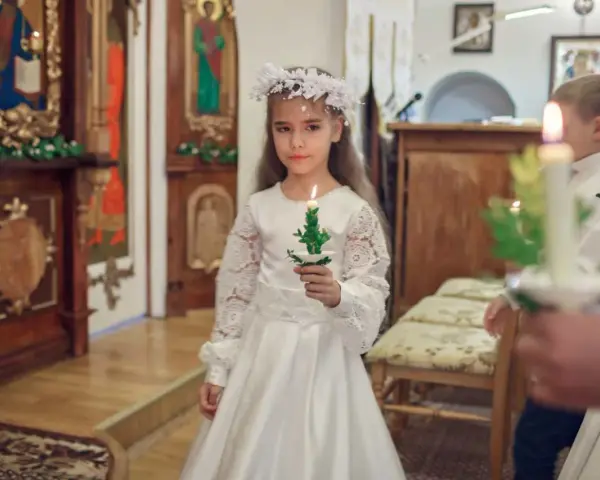 Dziewczynka w białej sukni i wianku trzyma zapaloną świecę. To ważny dzień, w której klasie komunia.