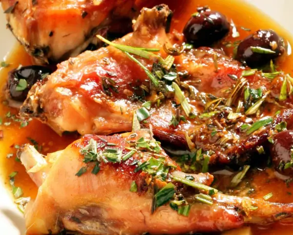 Coniglio alla ligure succulento, con olive nere e erbe aromatiche, servito in un piatto bianco.
