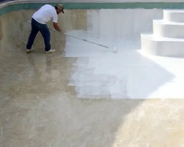 Hombre pintando una piscina de cemento con rodillo. La mitad de la piscina ya está pintada de blanco.