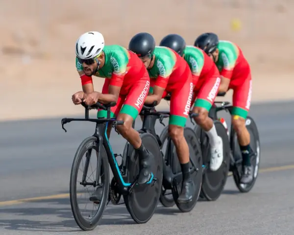 Équipe cycliste marocaine en tenue rouge et verte, concentrée sur la course. Ils roulent en peloton, tels des pur sang arabes noirs, sur un parcours désertique.