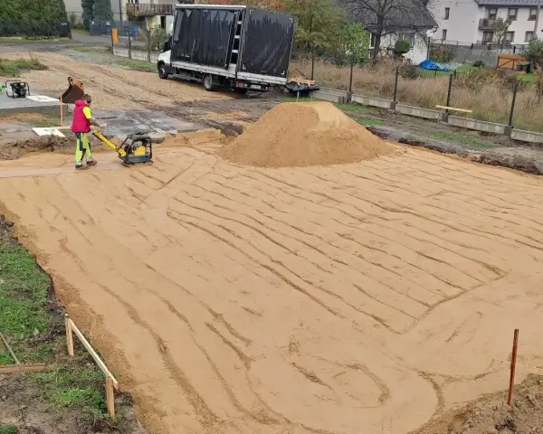 Pracownik zagęszcza piasek zagęszczarką. To kluczowy etap wymiany gruntu pod fundament, zapewniający stabilność przyszłego budynku.