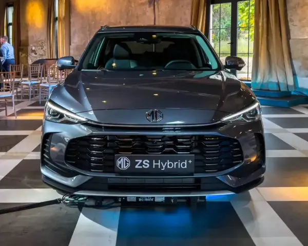 Szary MG ZS Hybrid+ prezentuje się elegancko na tle wnętrza z zasłonami i krzesłami.