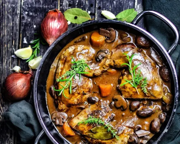 Herzhaftes Coq au Vin in einem Gusseisentopf, mit Pilzen, Karotten und Kräutern garniert. Daneben Zwiebeln und Knoblauch.