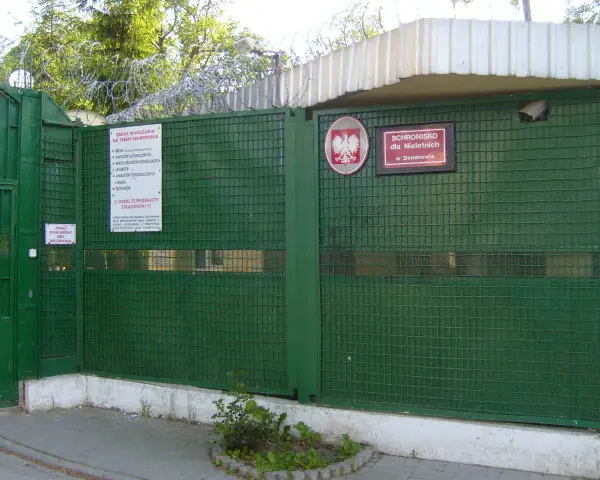 Zielona brama z drutem kolczastym i znakiem "Schronisko dla Nieletnich w Dominowie". To miejsce, gdzie młodzież potrzebuje wsparcia, a nie kary.