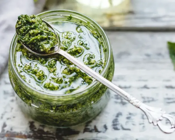Un vasetto di pesto genovese fatto in casa con frullatore, pronto per essere gustato. Un cucchiaino d'argento raccoglie un po' di questa delizia verde.
