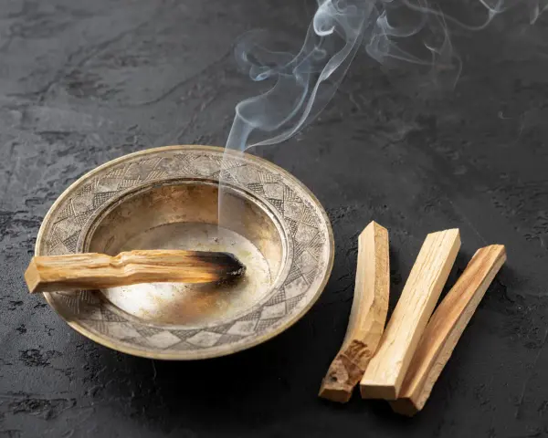 Un palo santo arde en un cuenco de metal, liberando humo blanco. A su lado, tres palos de palo santo sin quemar. Se puede quemar palo santo de noche para purificar el ambiente.