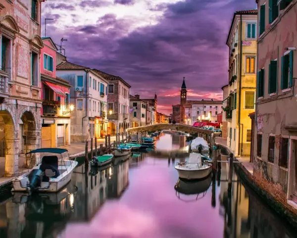 Un canale di Venezia al tramonto, con barche ormeggiate e riflessi viola sull'acqua. Un'atmosfera magica che ricorda i piombi di Venezia.