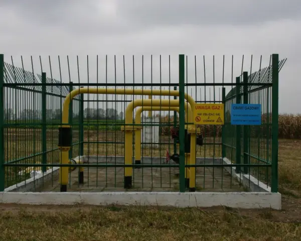 Żółty gazociąg wysokiego ciśnienia na działce, otoczony zielonym ogrodzeniem. Tablice ostrzegają przed gazem i ogniem.