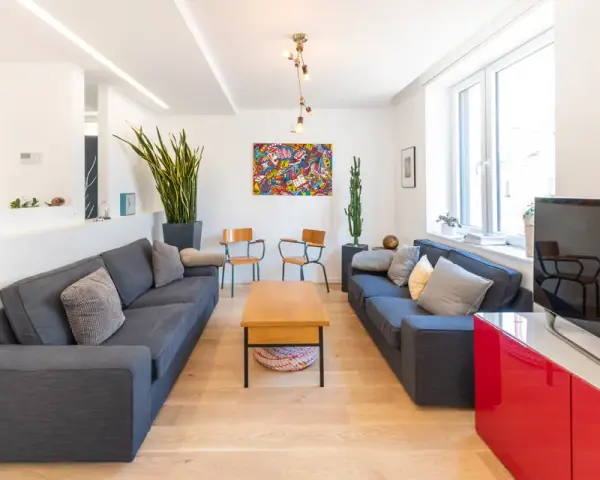 Soggiorno moderno e luminoso, ideale per mini appartamenti. Due divani grigi, tavolino in legno, quadri colorati e piante verdi.