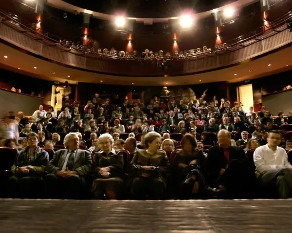 Widownia teatru, ludzie siedzą na miejscach, widać balkon z widzami.