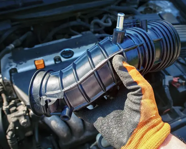 Gloved hand holds a black intake hose near a car engine. This part is often replaced during vacuum leak repair, affecting the vacuum leak repair cost.
