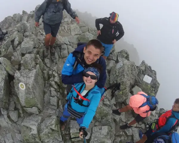 Grupa turystów na skalistym szczycie, otoczona mgłą. Jedna z kobiet, **mała wysoka**, robi sobie selfie z partnerem.