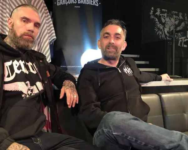 Deux hommes, dont un possible chanteur de Mass Hysteria, posent sur un canapé. L'un porte un sweat à capuche noir avec un aigle.