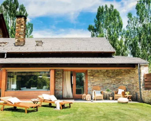 Fachadas de casas de pueblo modernas con piedra y madera, jardín con tumbonas y zona de estar exterior.