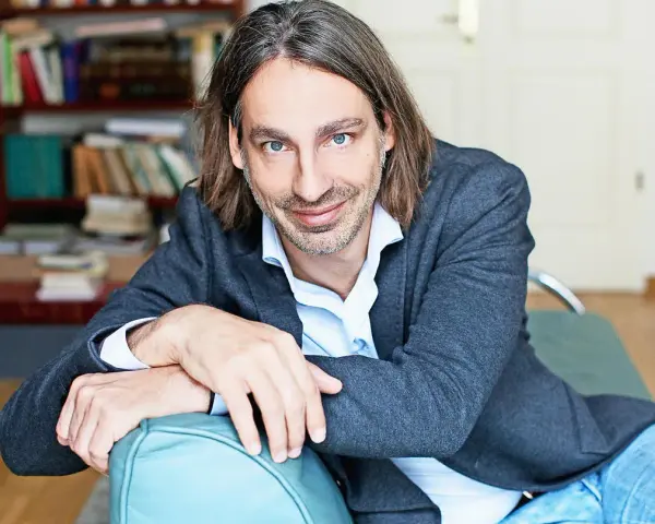 Richard David Precht sitzt lächelnd auf einem türkisfarbenen Sofa, im Hintergrund ein Bücherregal.