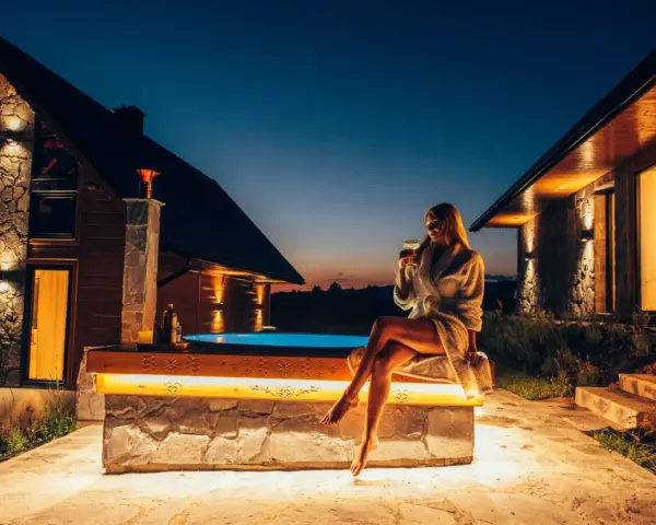Kobieta w szlafroku z drinkiem siedzi przy jacuzzi przed górskim domkiem.