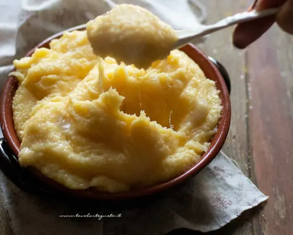 Un cucchiaio solleva una porzione filante di polenta concia valdostana, calda e cremosa, in una ciotola di terracotta.