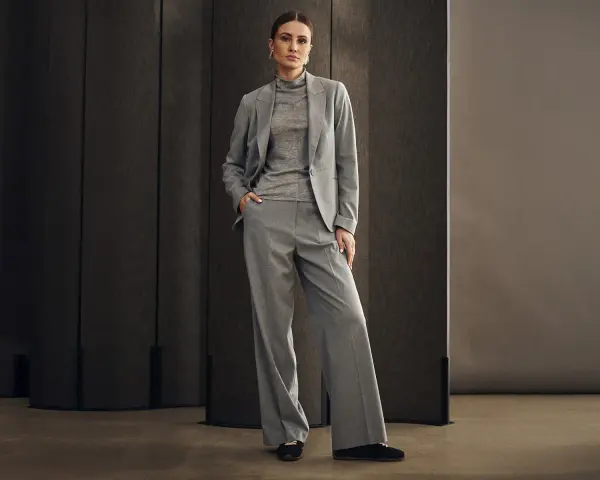 Elegancka kobieta w szarym garniturze, idealnym na wesele. Spodnie z szerokimi nogawkami i marynarka tworzą stylowy zestaw.