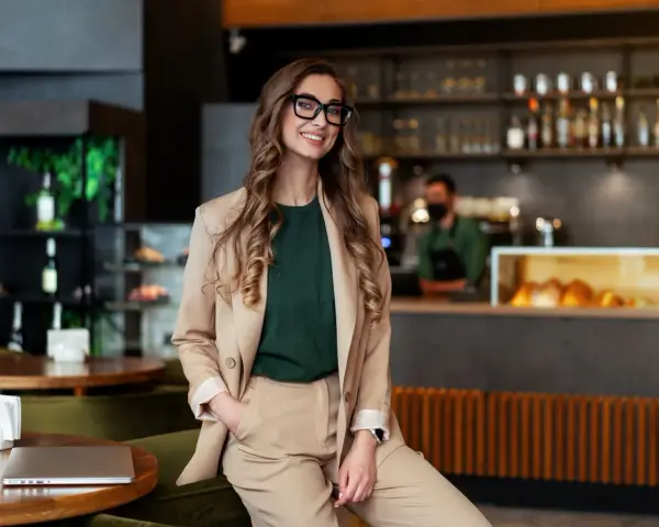 Mujer sonriente con gafas, vestida de traje beige, sentada en un restaurante moderno. Detrás, un barista trabaja. Esencial para la gestión de restaurantes.
