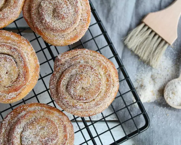 Nybakade karlsbaderbullar med socker och kanel på ett svart galler. En borste och en sked med socker ligger bredvid.