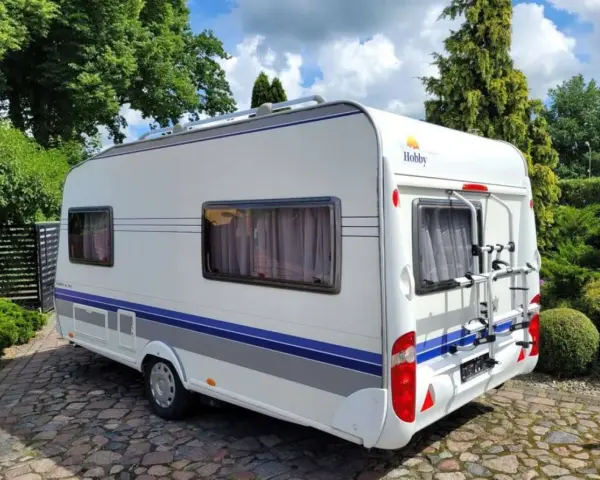 Biała przyczepa kempingowa Hobby 440 z niebieskimi pasami, zaparkowana na brukowanym podjeździe. Idealna do podróżniczego hobby.