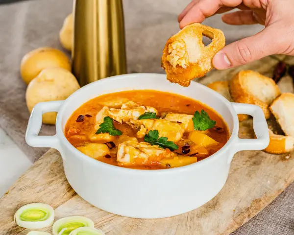 Una mano intinge un crostino nella zuppa di pesce surgelato, ricca di sapori e profumi.