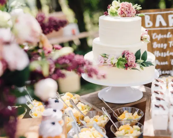 Elegancki tort weselny boho ozdobiony kwiatami, obok deserki i owoce.
