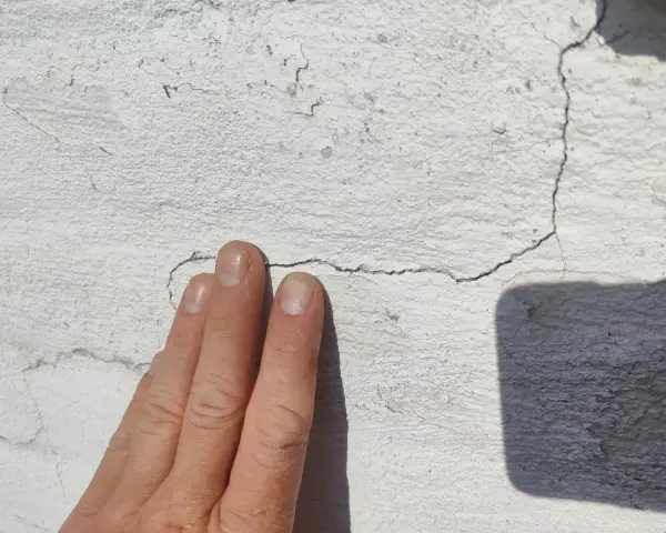 En hand pekar på sprickor i en vit, texturerad fasad.