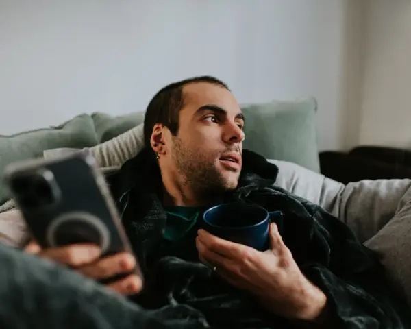 Mann liegt auf Sofa, hält Tasse und Handy. Mögliche Bewegungsmangel Symptome wie Müdigkeit und Lethargie werden angedeutet.