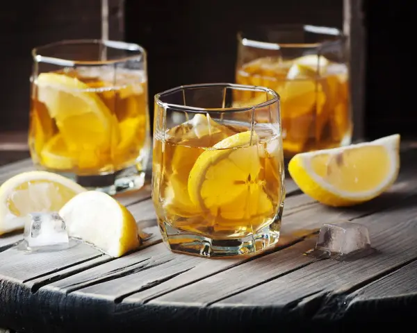 Metaxa orange z czym pić? Idealnie z lodem i plasterkiem cytryny. Trzy szklanki złocistego trunku na drewnianym blacie.