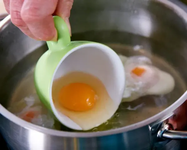 Ein rohes Ei wird aus einer grünen Tasse in kochendes Wasser gegossen, um verlorene Eier zuzubereiten.