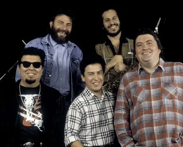 Los Lobos, native sons, pose together. One man wears sunglasses and a fringed jacket, another a denim jacket, and two sport plaid shirts. A saxophone is visible.