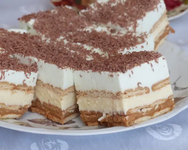 Szybkie ciasto bez pieczenia z budyniem i herbatnikami, przełożone kremem karmelowym, posypane wiórkami czekolady.