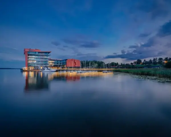 Nowoczesny hotel na ptasiej wyspie, otoczony spokojną wodą i zielenią, z łodziami przy pomoście.