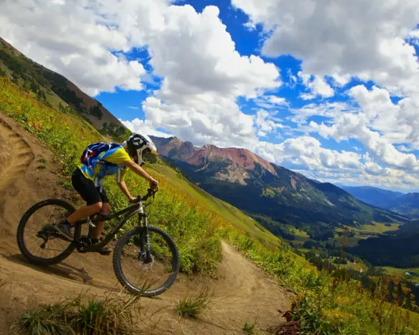 A mountain biker navigates a steep, winding trail, experiencing the best mountain biking in Colorado with stunning valley views and dramatic clouds.