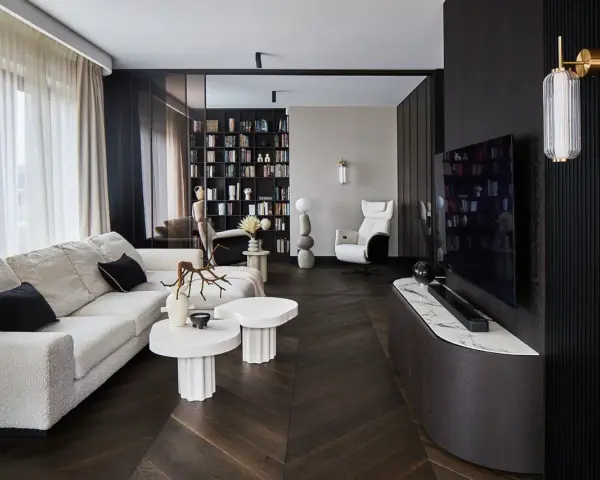 Nowoczesny salon z wygodną sofą, stolikami i regałem pełnym książek. Dominują ciemne, grafitowe akcenty, tworząc elegancką atmosferę.