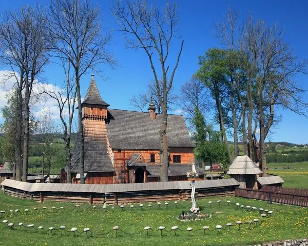 Drewniany kościół w Dębnie, otoczony kamiennym murem i drzewami, z krzyżem misyjnym na trawiastym dziedzińcu.